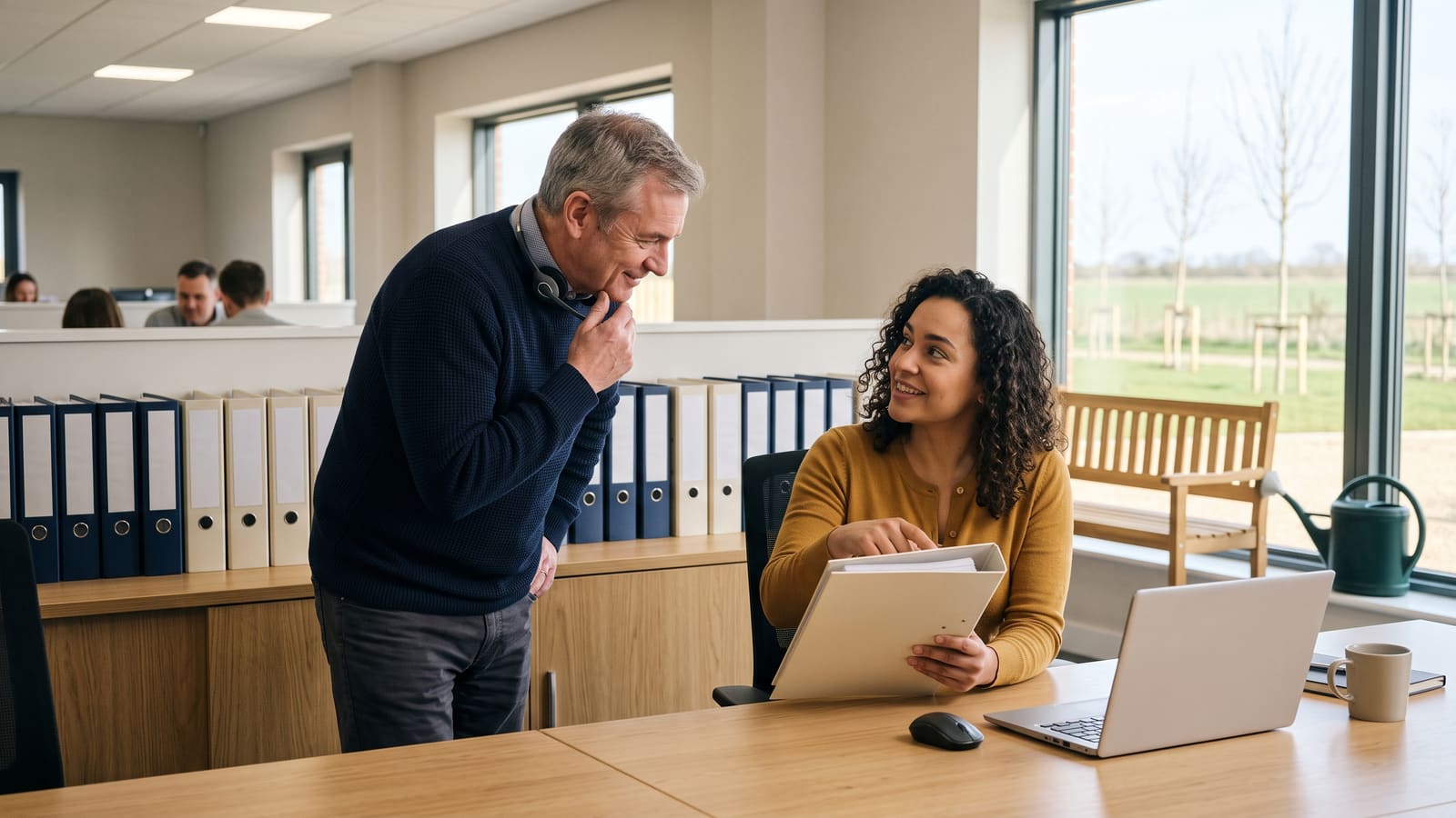 A customer service lead in a navy jumper with a headset round his neck asks a smiling colleague a question as she checks a product binder, in a bright business-park office with shelves of blank-spined binders